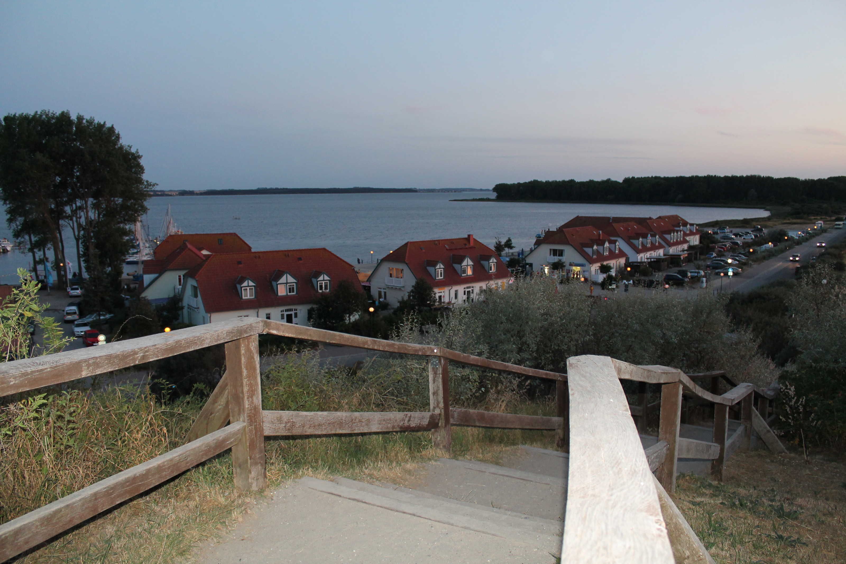 Eine Küstenstadt mit Holzbühnen, die zu einem Strand führen, Häuser mit roten Dächern und einem ruhigen Meer.