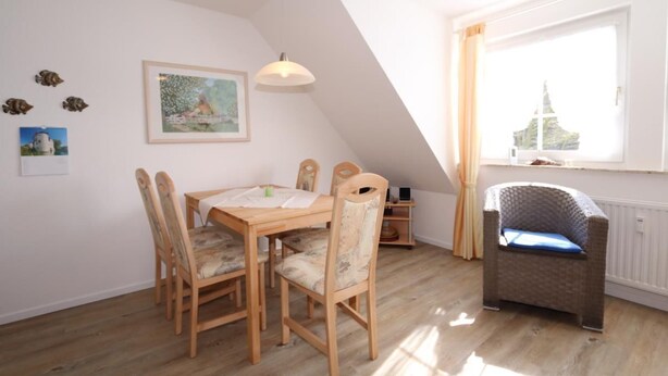 A dining area with a wooden table and chairs, a pendant light, a framed picture on the wall, and a window with curtains. - Vacation apartment Reinhardt (Sylt-Ost)