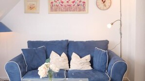 A blue sofa with white pillows, a glass coffee table with a vase of flowers, a framed painting, a clock, and a floor lamp. - Vacation apartment Reinhardt (Sylt-Ost)