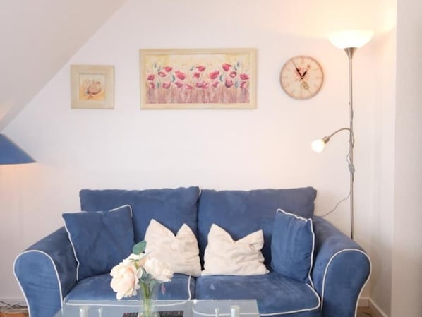 Un canapé bleu avec des coussins blancs, une table basse en verre ornée d’un vase de fleurs, un tableau encadré, une horloge et une lampe sur pied.
