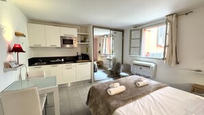 Ein kleines Hotelzimmer mit Küchenzeile, einem Bett mit brauner Decke, einem Schreibtisch mit Stuhl und einem Fenster mit Vorhängen.