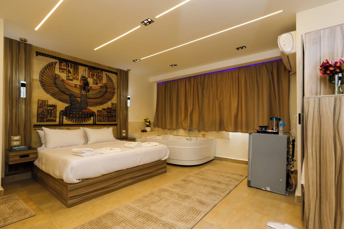 Una habitación de hotel moderna con una cama grande, una zona de estar con una mesita y sillas, un gran elemento decorativo en la pared, un amplio ventanal con cortinas y un refrigerador.