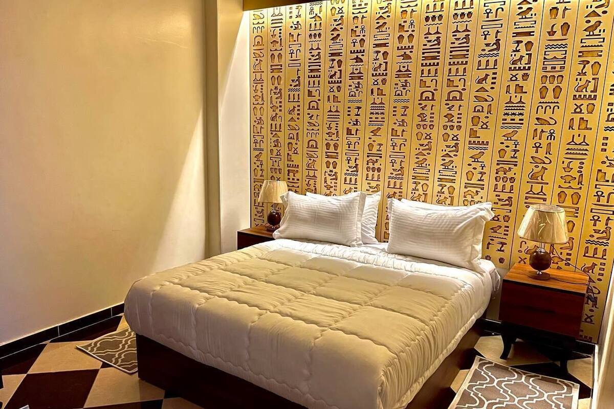 Una habitación de hotel con cama, mesitas de noche y una pared con jeroglíficos.