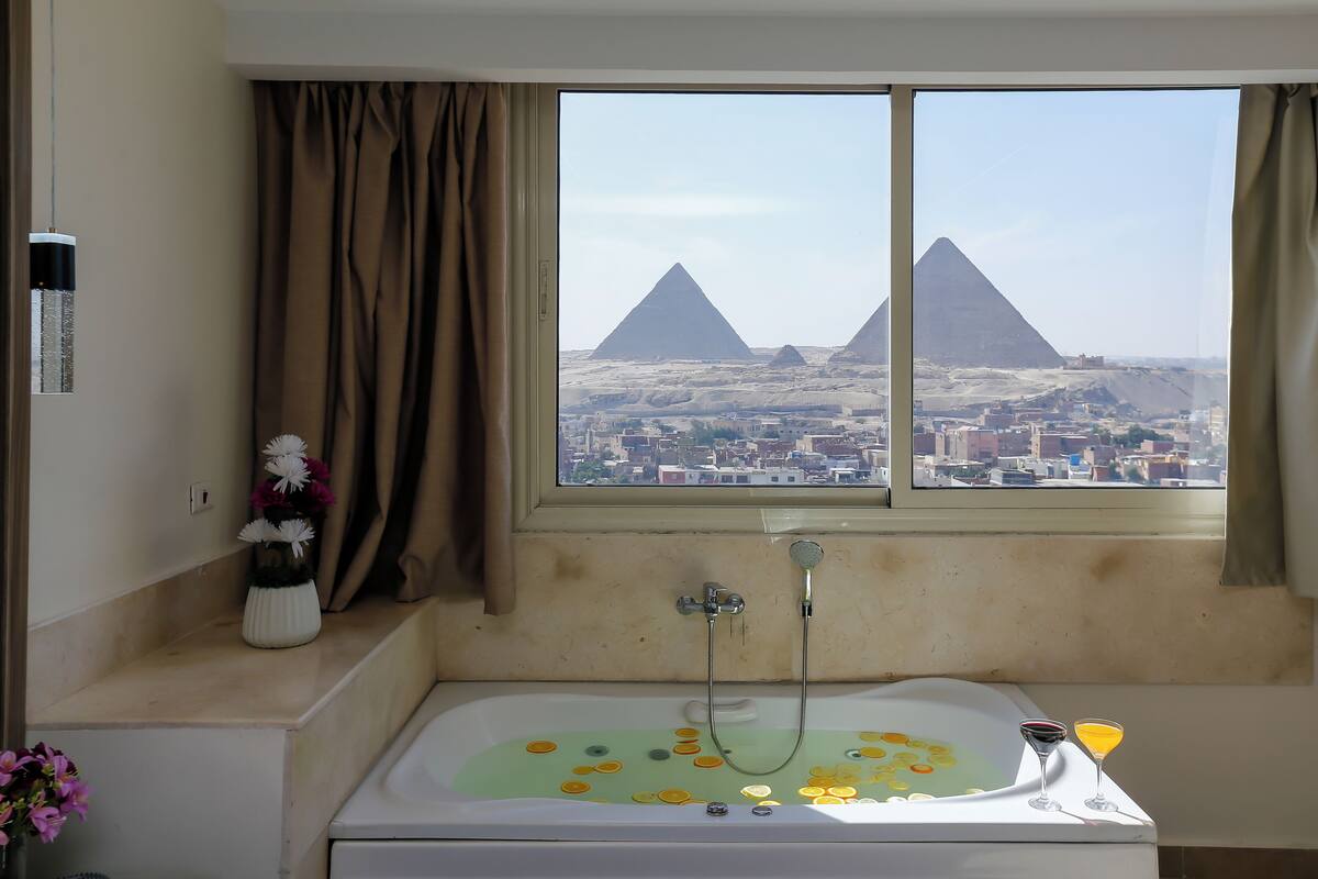 Un baño con bañera, una ventana con vistas a pirámides y un florero con flores en el alféizar.