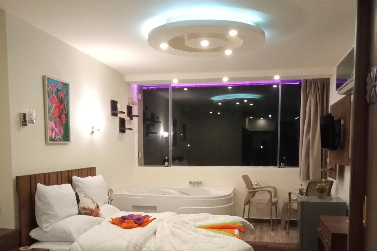 Una habitación de hotel moderna con una cama grande, un escritorio, una silla, una mesita pequeña, una bañera y una ventana con vistas a la ciudad de noche.