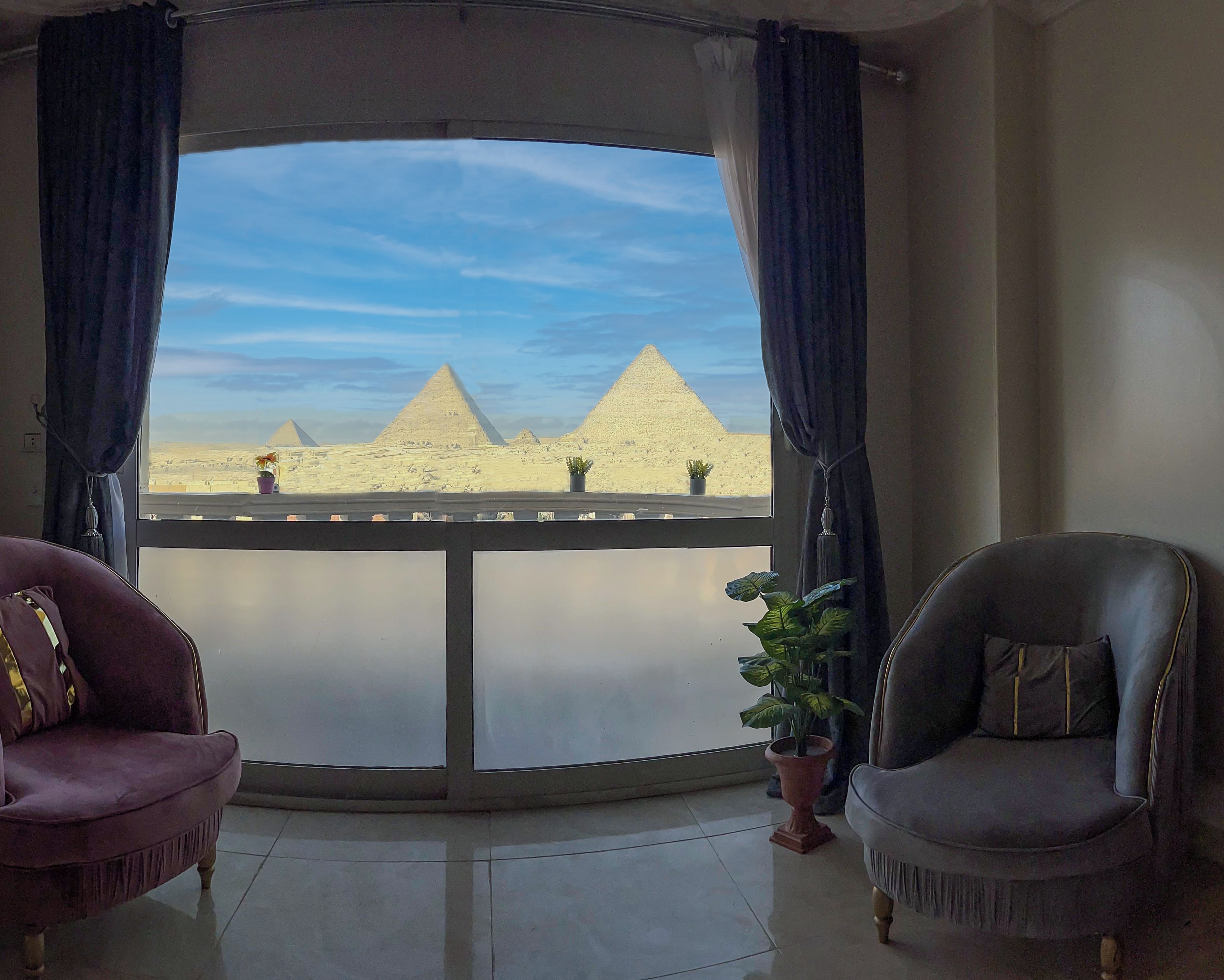 A room with a view of the pyramids, a purple armchair, a grey armchair, and a potted plant.