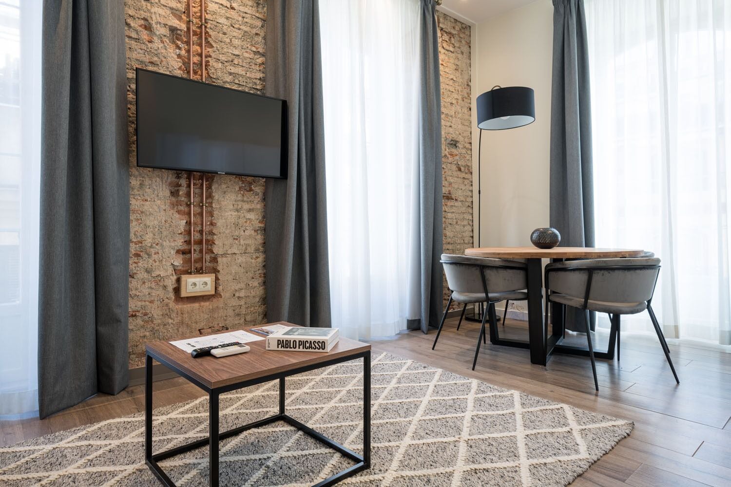 A modern living room with a brick wall, a flat-screen TV, a wooden coffee table with a book titled "PABLO PICASSO", a dining table with chairs, and a floor lamp.
