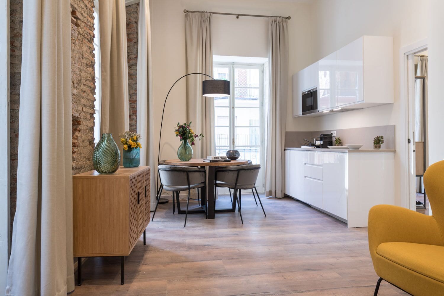 A modern kitchen with white cabinets, a dining area with a wooden table and chairs, and a yellow armchair.