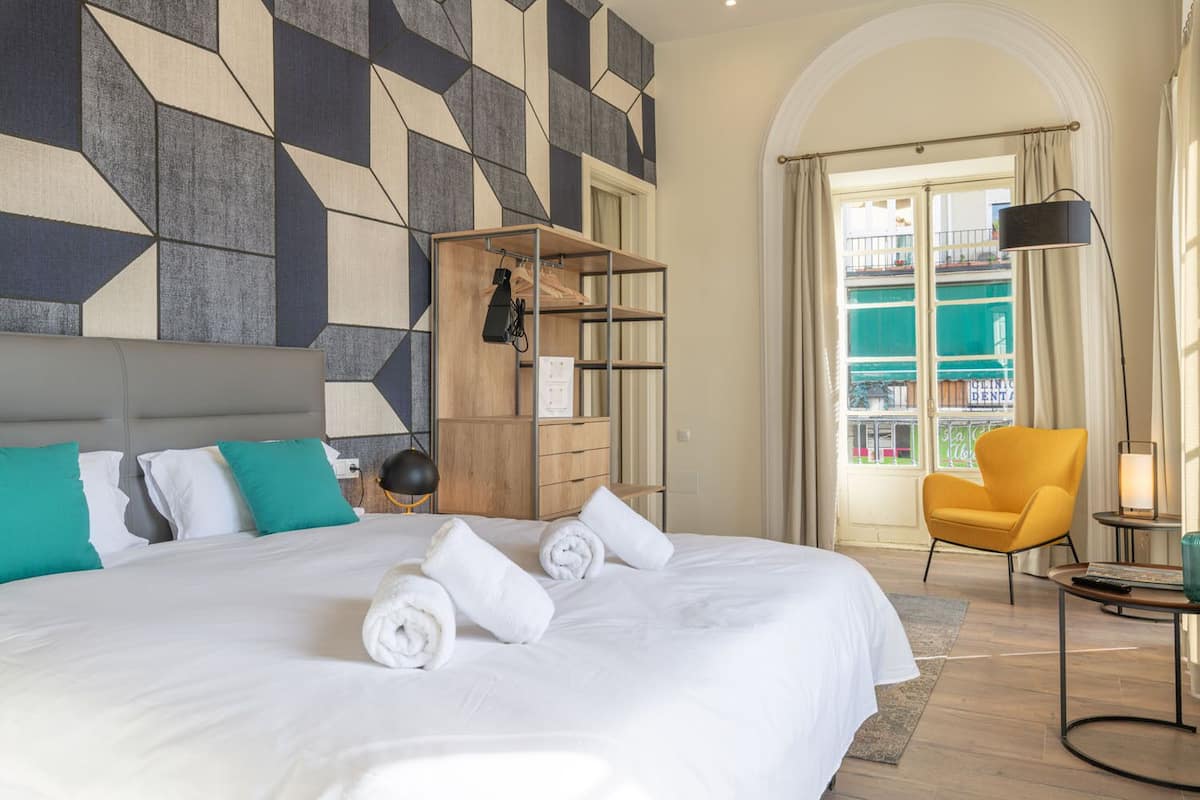 A hotel room with a large bed, a wooden wardrobe, a yellow armchair, and a view of a balcony with a pool.