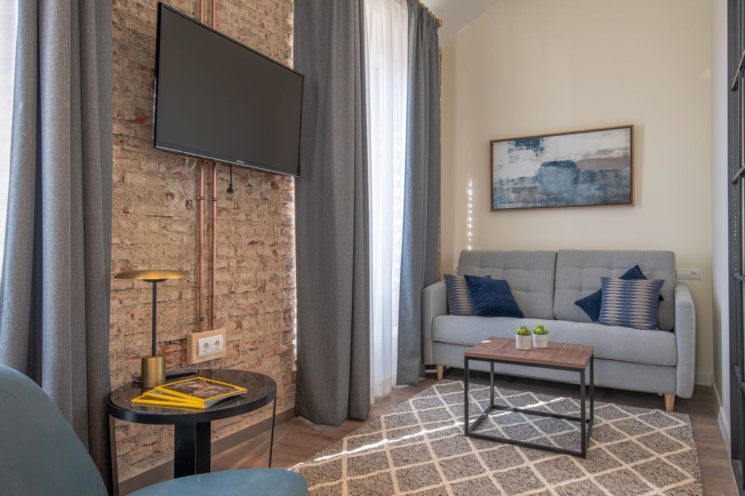 Una sala de estar con un sofá gris, un televisor de pantalla plana montado en una pared de ladrillo, una mesita redonda con una lámpara y una alfombra estampada.