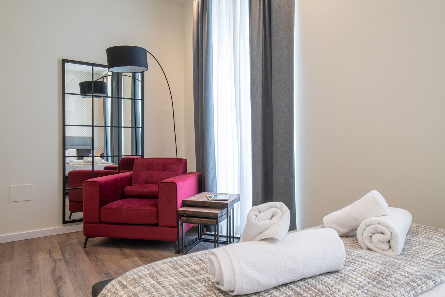 A hotel room with a red armchair, a small table, a bed with rolled towels, a floor lamp, and a large mirror.