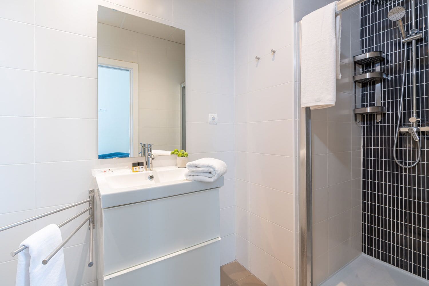 Un baño moderno con lavamanos blanco, espejo y ducha con pared de azulejos negros.