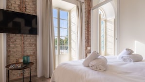 A hotel room with a brick wall, a large bed with white linens, a TV mounted on the wall, and a small table with a magazine and a green vase. - Homeart Apartments Malaga (Málaga)