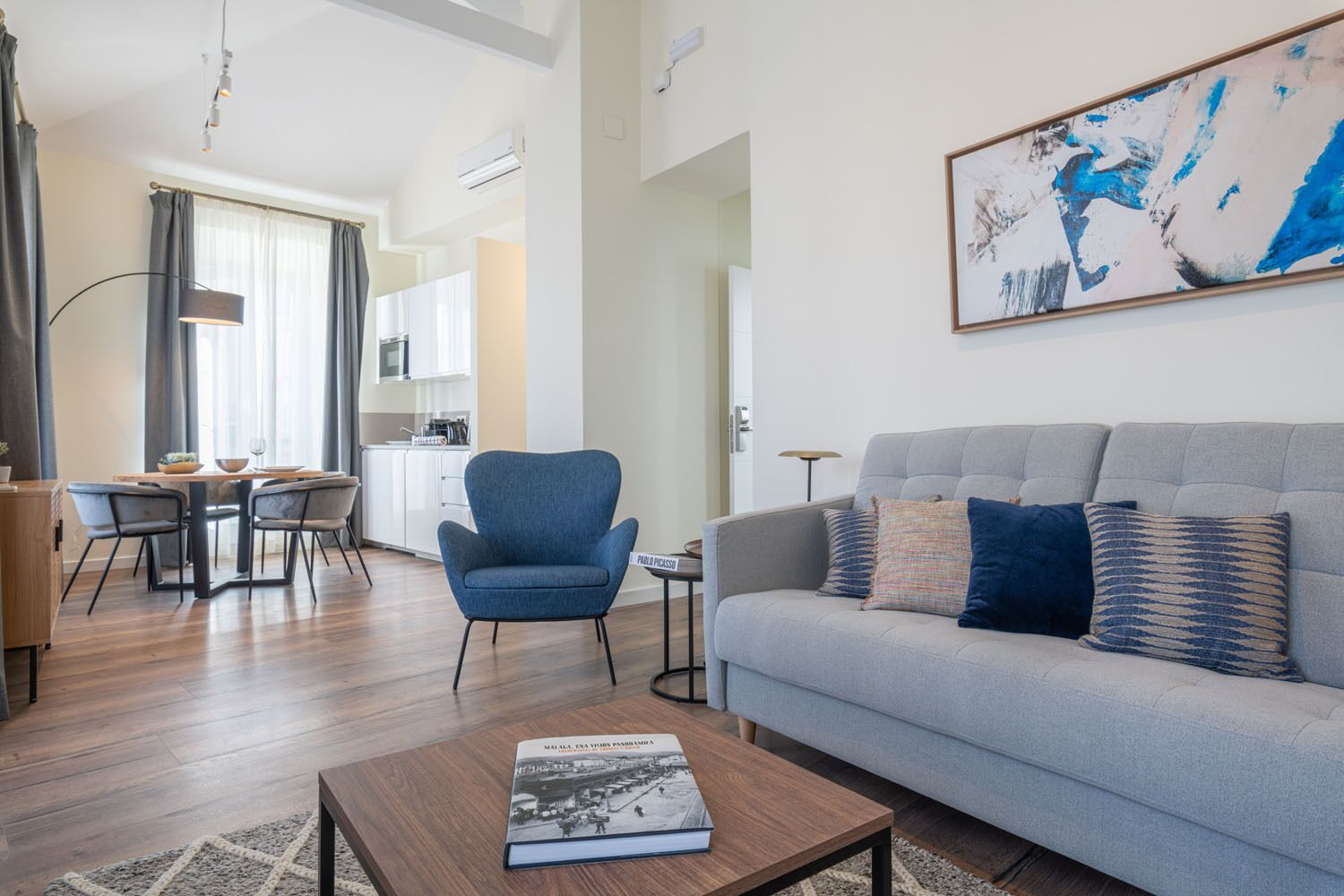 A modern living room with a grey sofa, a blue armchair, and a coffee table with a book. There is a dining area with a table and chairs, and a kitchen in the background.