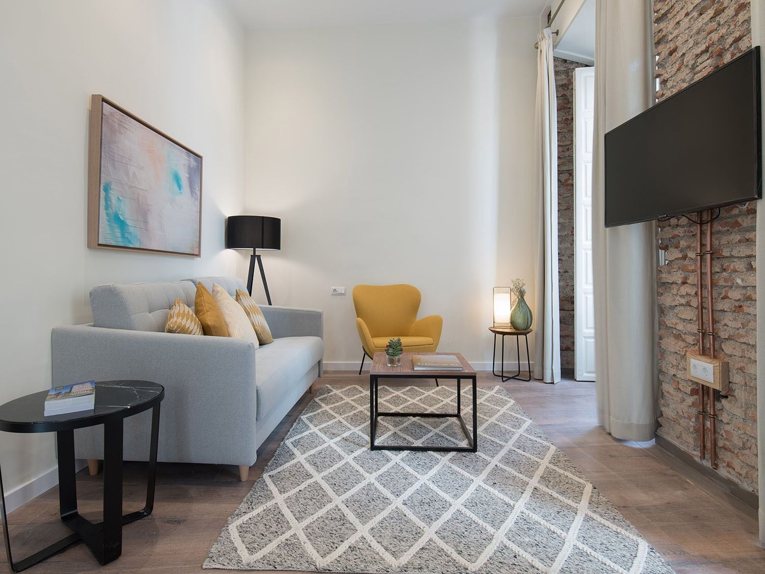 Un salon moderne avec un canapé gris, un fauteuil jaune, une table basse et un téléviseur fixé à un mur en briques.