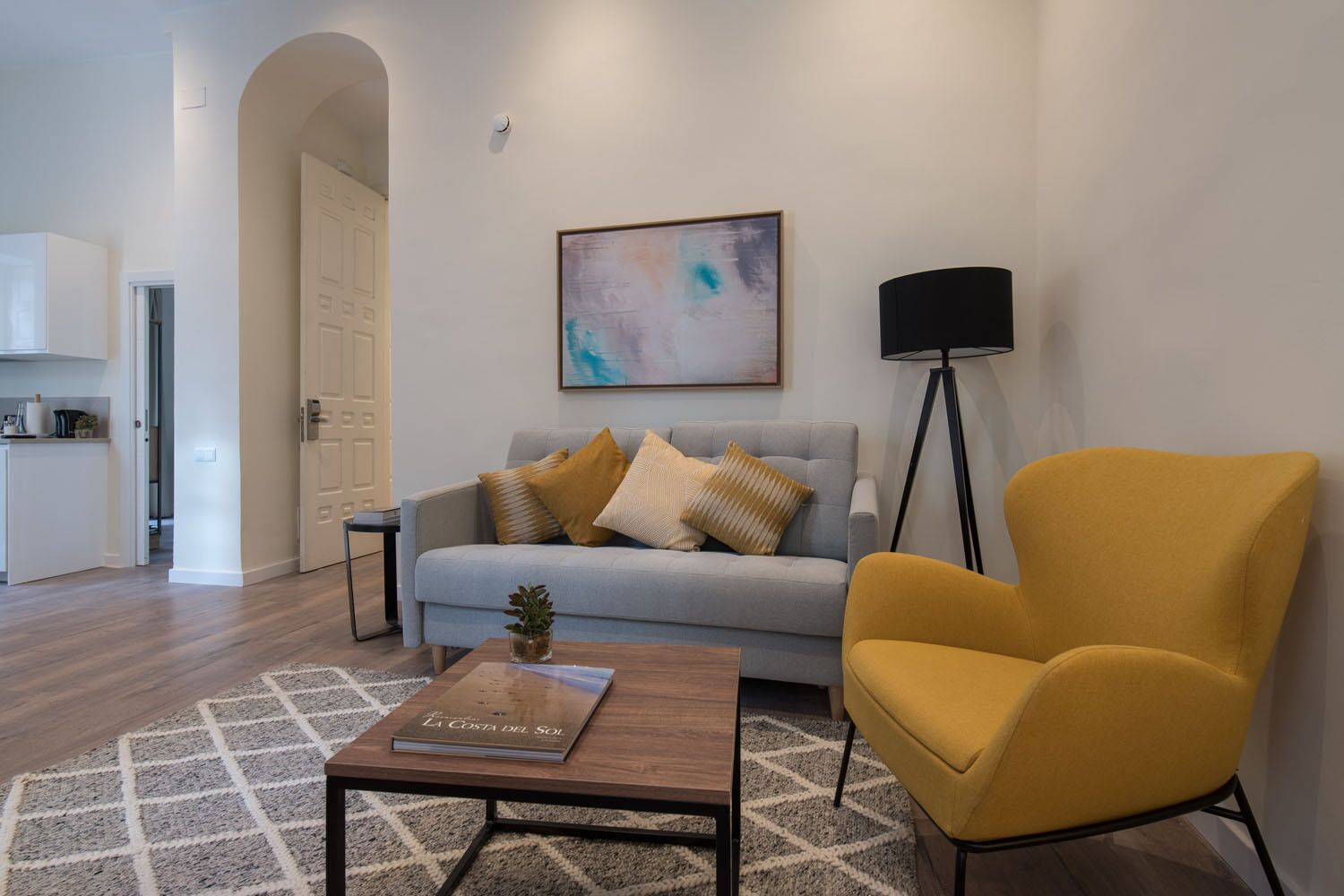 A modern living room with a sofa, a yellow armchair, a coffee table with a book, a floor lamp, and a painting on the wall.