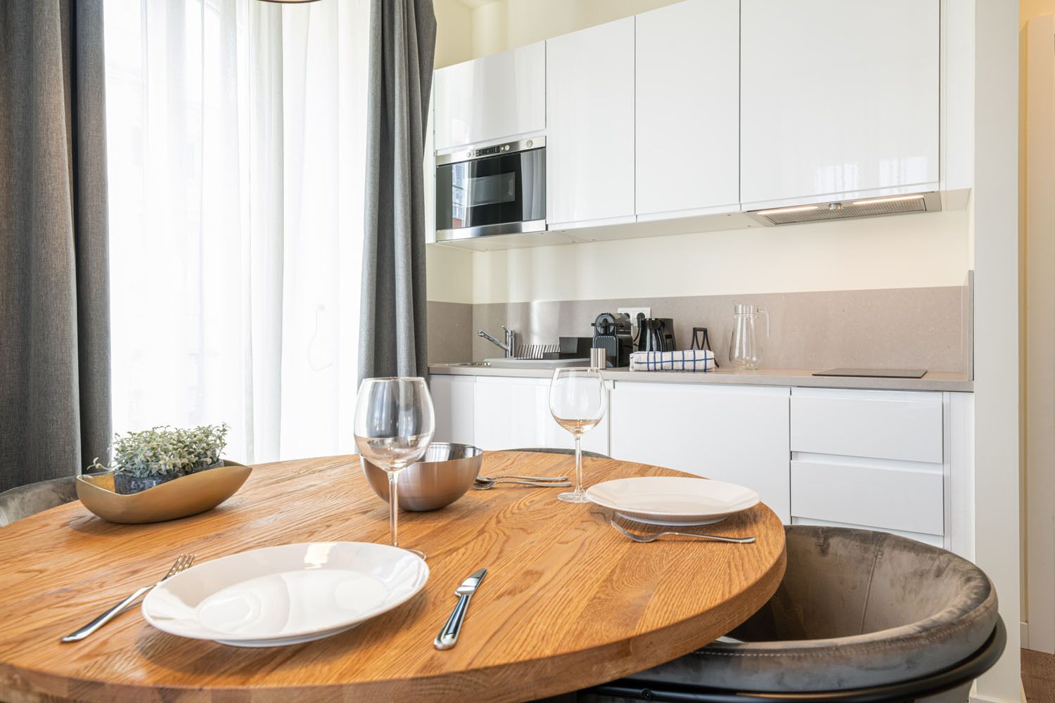 A modern kitchen with white cabinets, a microwave, and a sink. There is a wooden dining table with plates, cutlery, and a wine glass.