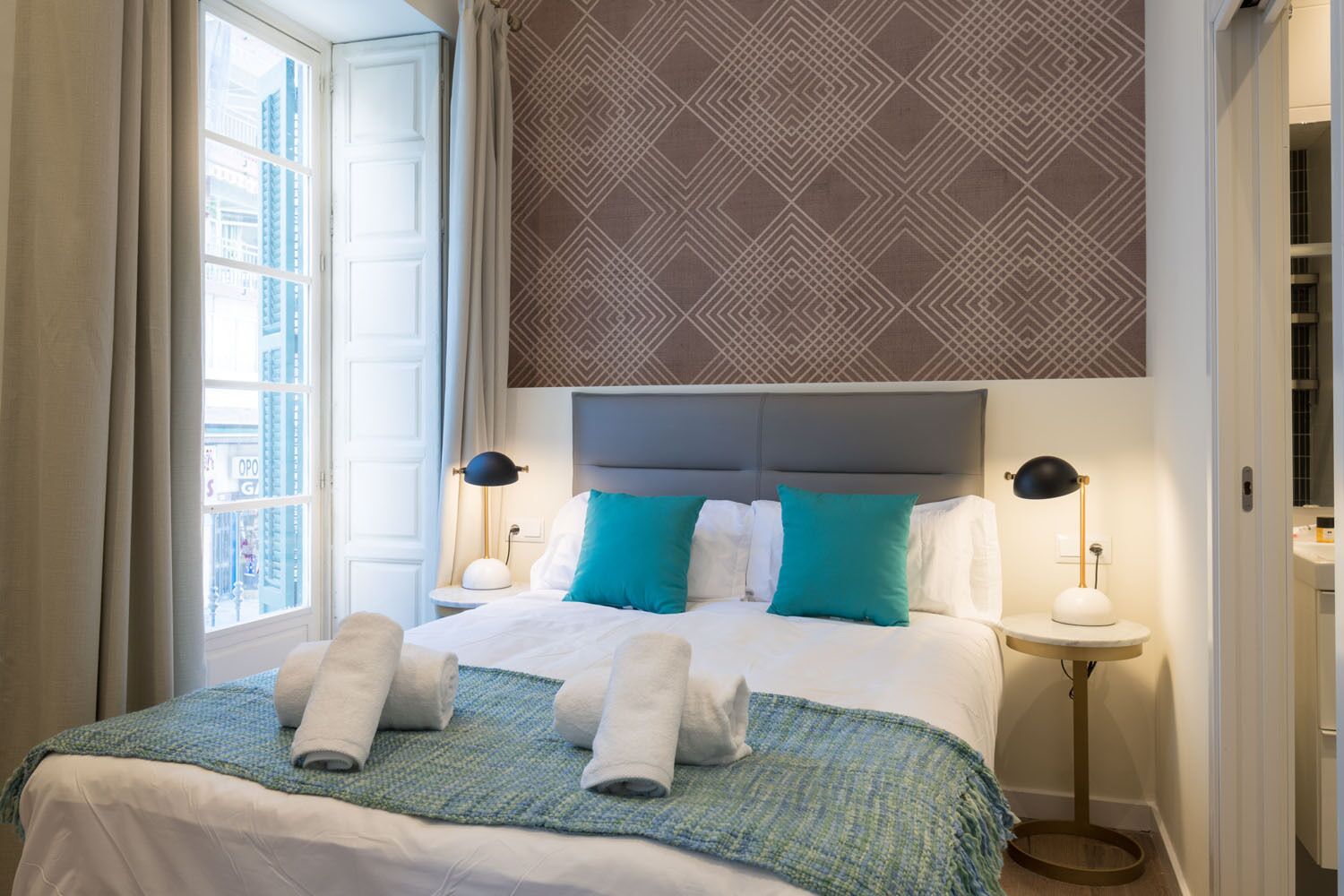 Habitación de hotel con una cama grande, dos lámparas de noche, un cabecero estampado y vistas de la ciudad a través de una ventana.