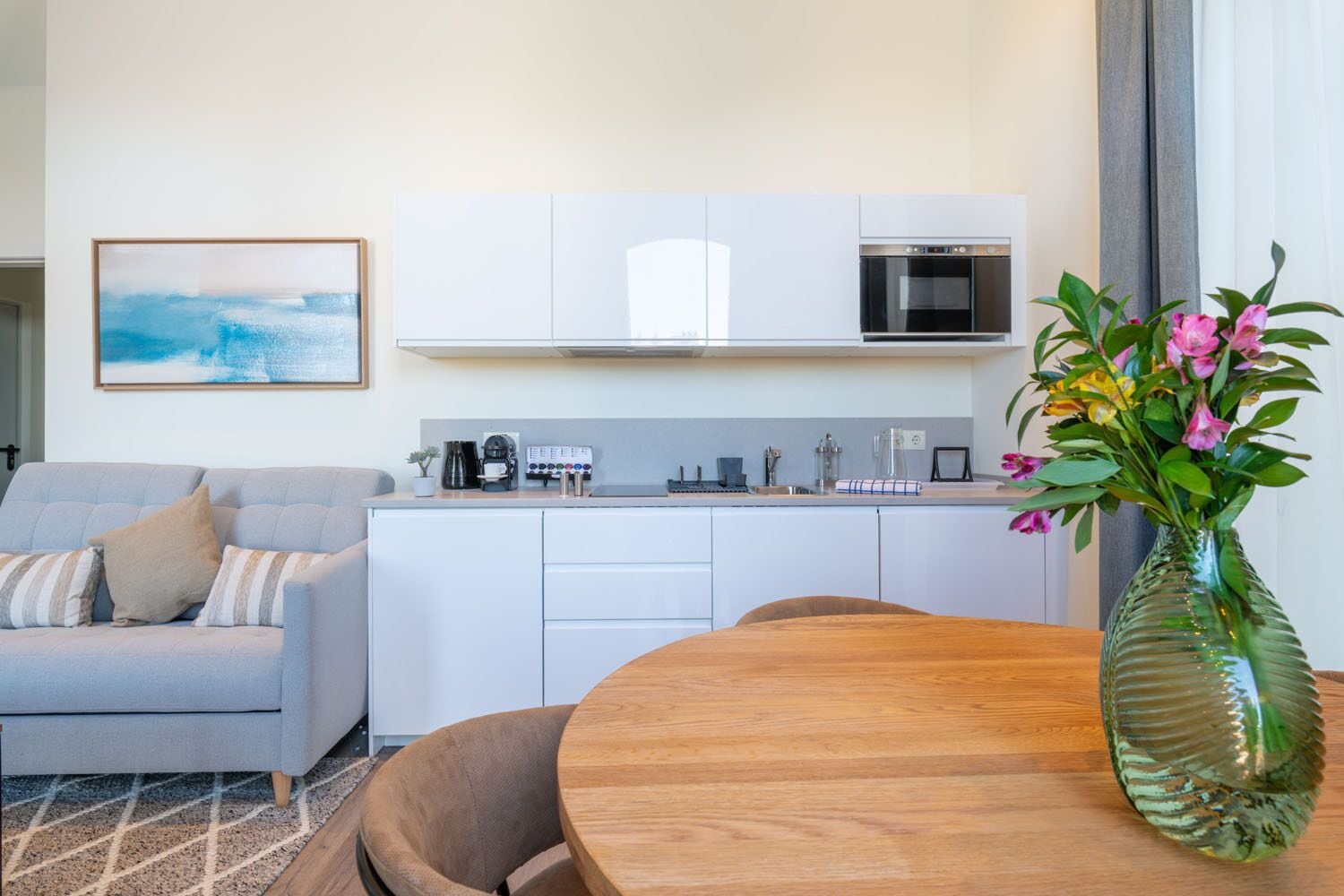 Une cuisine moderne avec des meubles blancs, un four encastré et une machine à café. On y trouve une table à manger avec des chaises et un vase de fleurs.