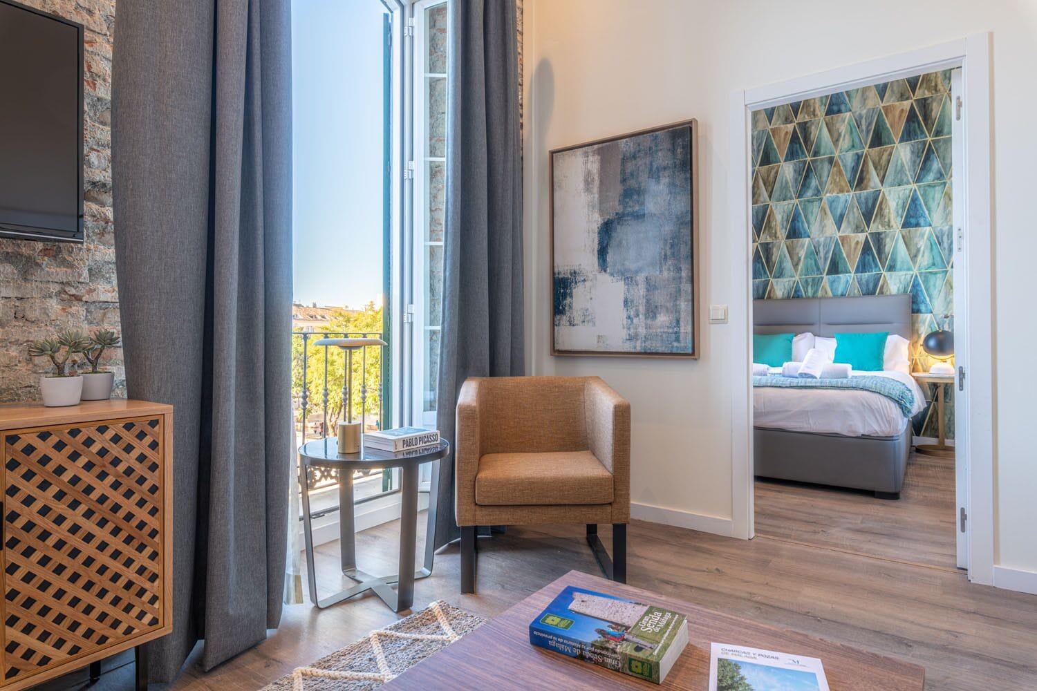 Een moderne hotelkamer met een groot raam, een comfortabele fauteuil, een tafeltje met een boek en een bed met een hoofdeinde met een geometrisch patroon.
