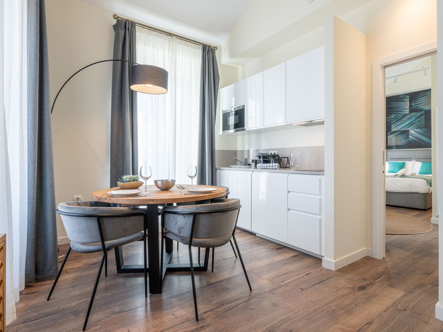 A modern kitchen with white cabinets, a round dining table with four chairs, and a large window with grey curtains.