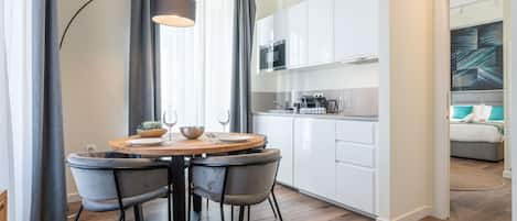 A modern kitchen with white cabinets, a round dining table with four chairs, and a large window with grey curtains.