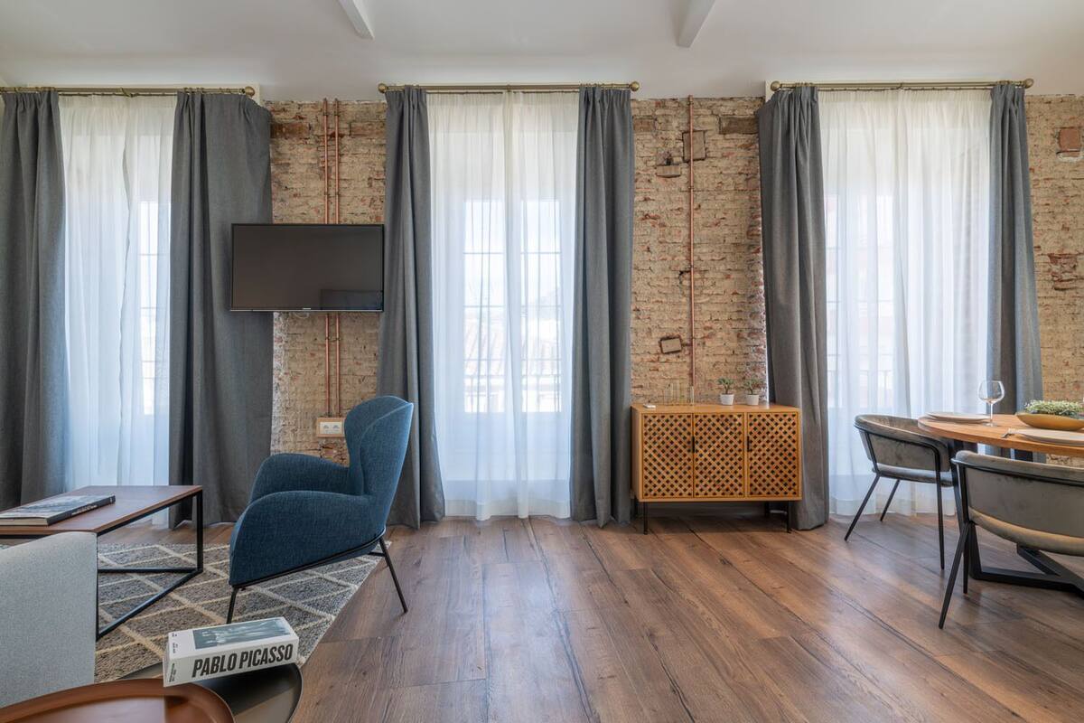 A modern living room with exposed brick walls, a flat-screen TV mounted on the wall, a blue armchair, a wooden dining table with chairs, and a small cabinet with a plant.