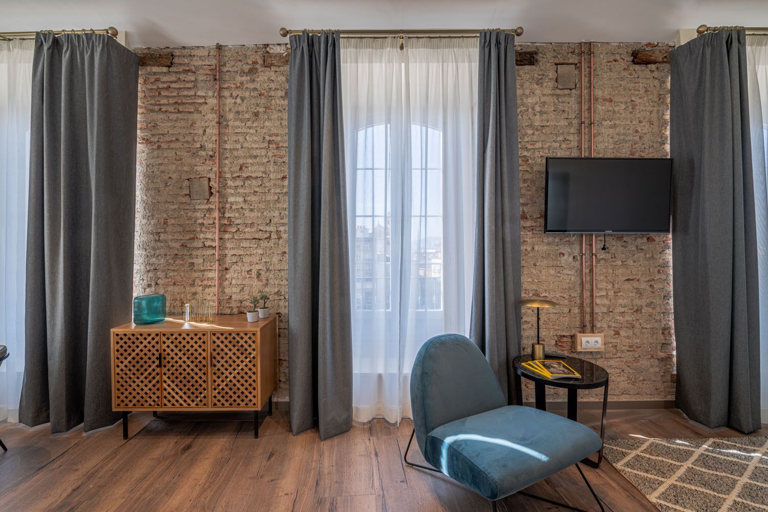 A room with a brick wall, a wooden cabinet, a blue chair, a small table with a lamp, and a TV mounted on the wall.