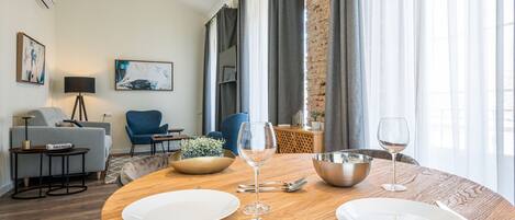 A dining area with a round wooden table set for two, two wine glasses, a bowl, and cutlery. There is a sofa and a chair in the background, a lamp, and a painting on the wall.