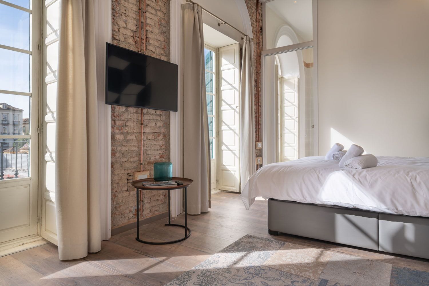 Un dormitorio moderno con una cama grande, un televisor de pantalla plana montado en una pared de ladrillo y vistas a la ciudad a través de amplias ventanas.