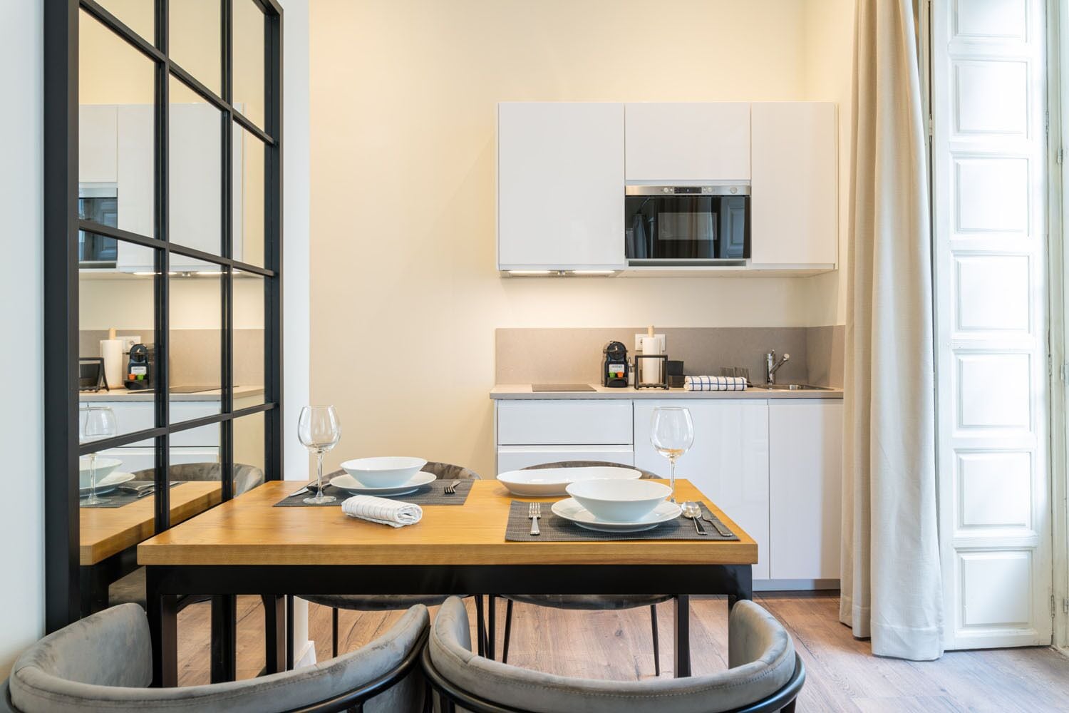 A modern kitchen with white cabinets, a microwave, and a coffee machine. There is a dining table with chairs and a glass of wine.
