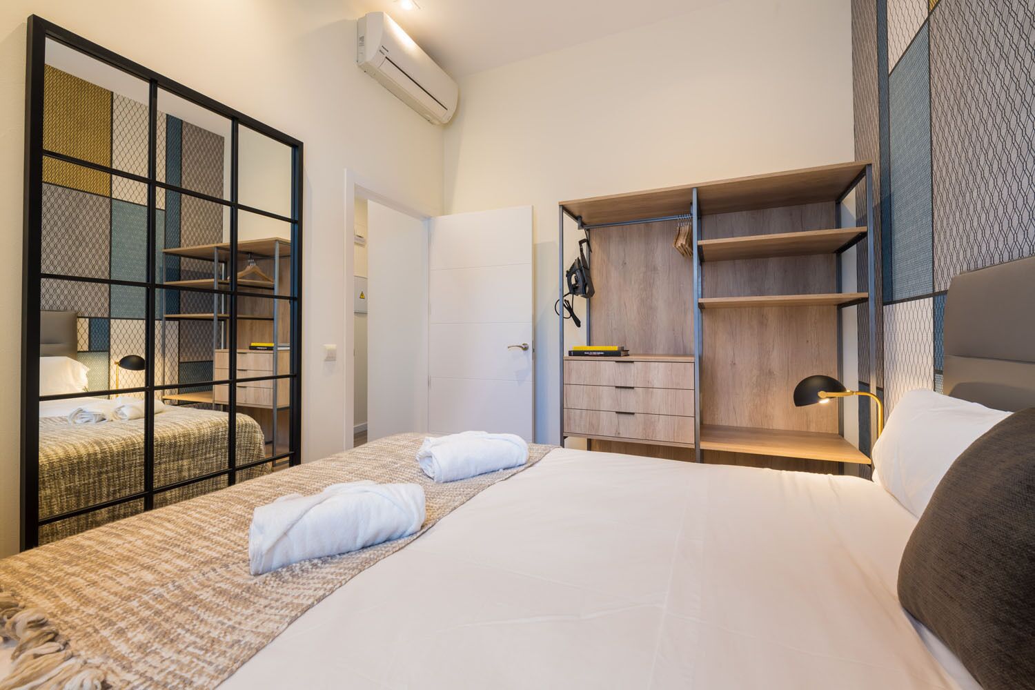 A modern hotel room with a large bed, a wardrobe with hanging space, and a built-in desk with a lamp.