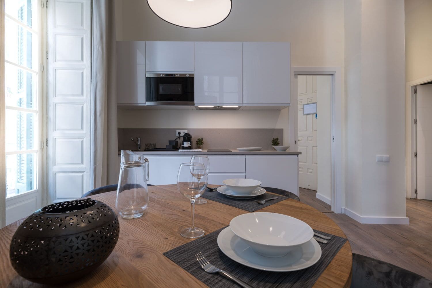 A modern kitchen with white cabinets, a microwave, and a coffee machine. There is a dining table with a pitcher, wine glasses, and plates.