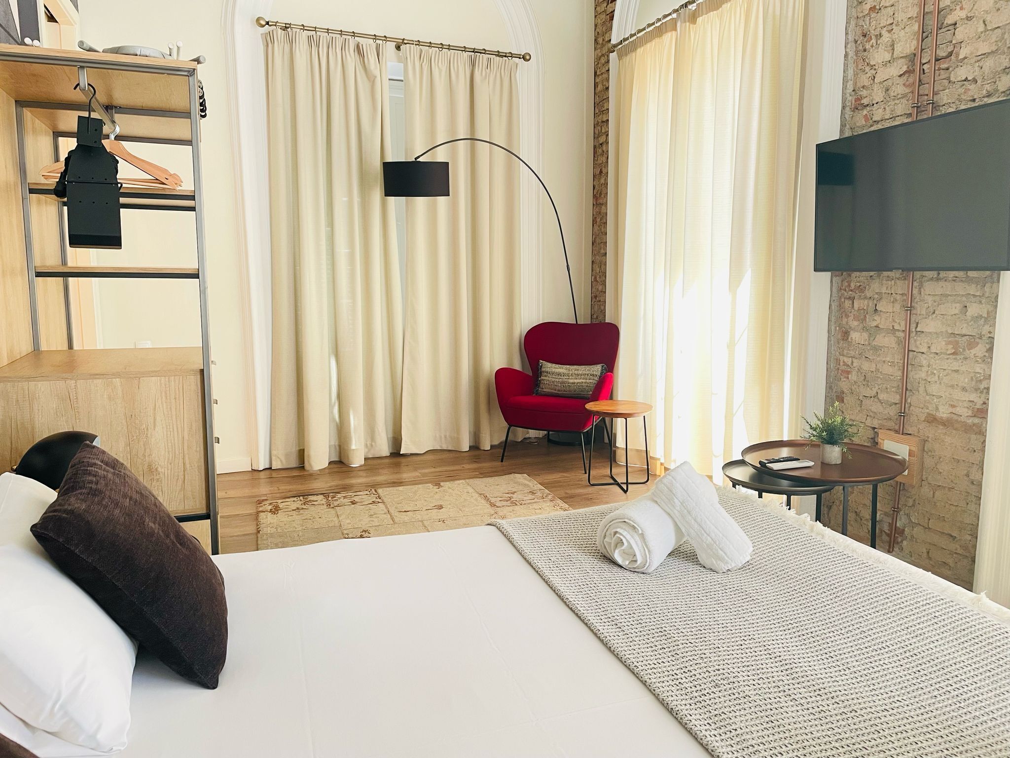 Een hotelkamer met een bed, een rode fauteuil, een klein tafeltje, een televisie en een houten plank met kleding.