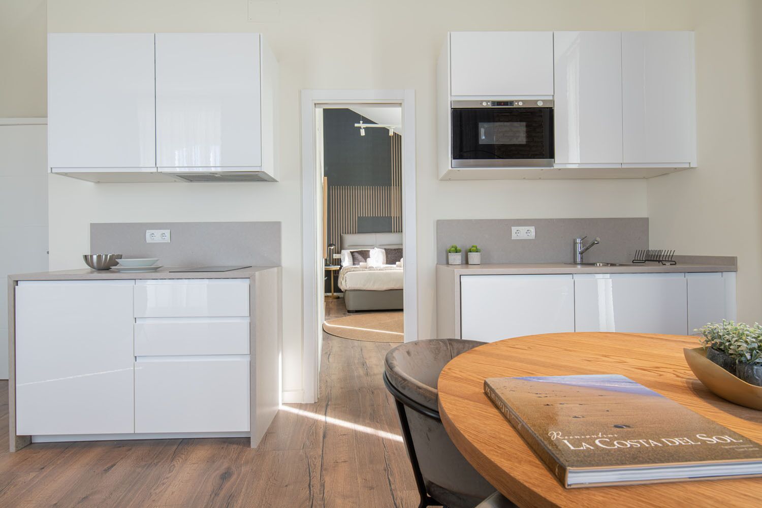 A modern kitchen with white cabinetry, a built-in oven, and a dining area with a wooden table and a book titled "La Costa del Sol".