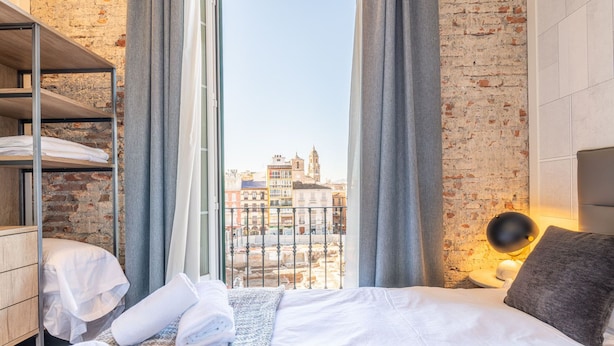 A hotel room with a bed, a wooden wardrobe, a window with curtains, and a view of buildings. - Homeart Apartments Malaga (Málaga)