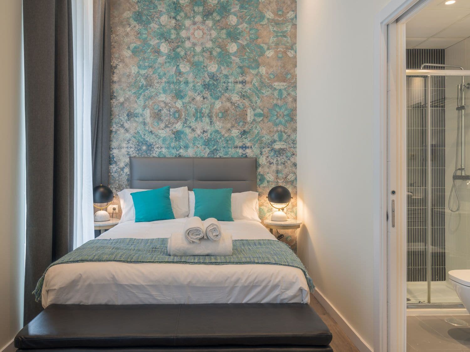 A modern hotel room with a large bed, a bench, a nightstand with a lamp, and a shower area visible through a glass door.