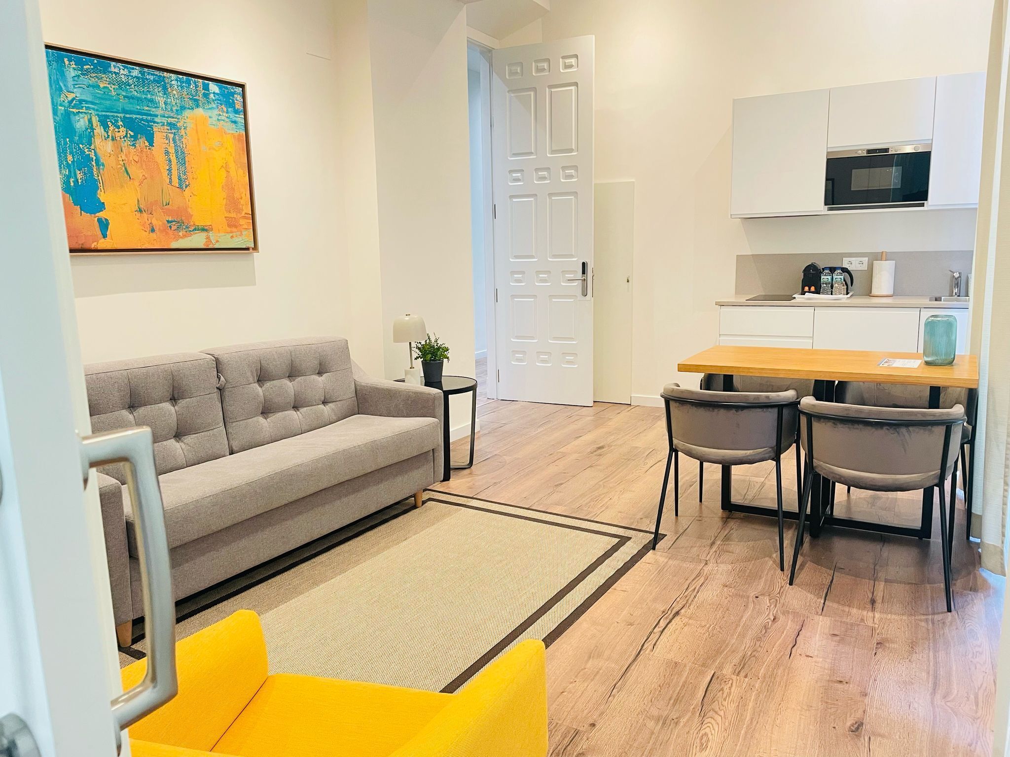 A modern apartment with a grey sofa, yellow armchair, and a kitchen area with white cabinets and a microwave.