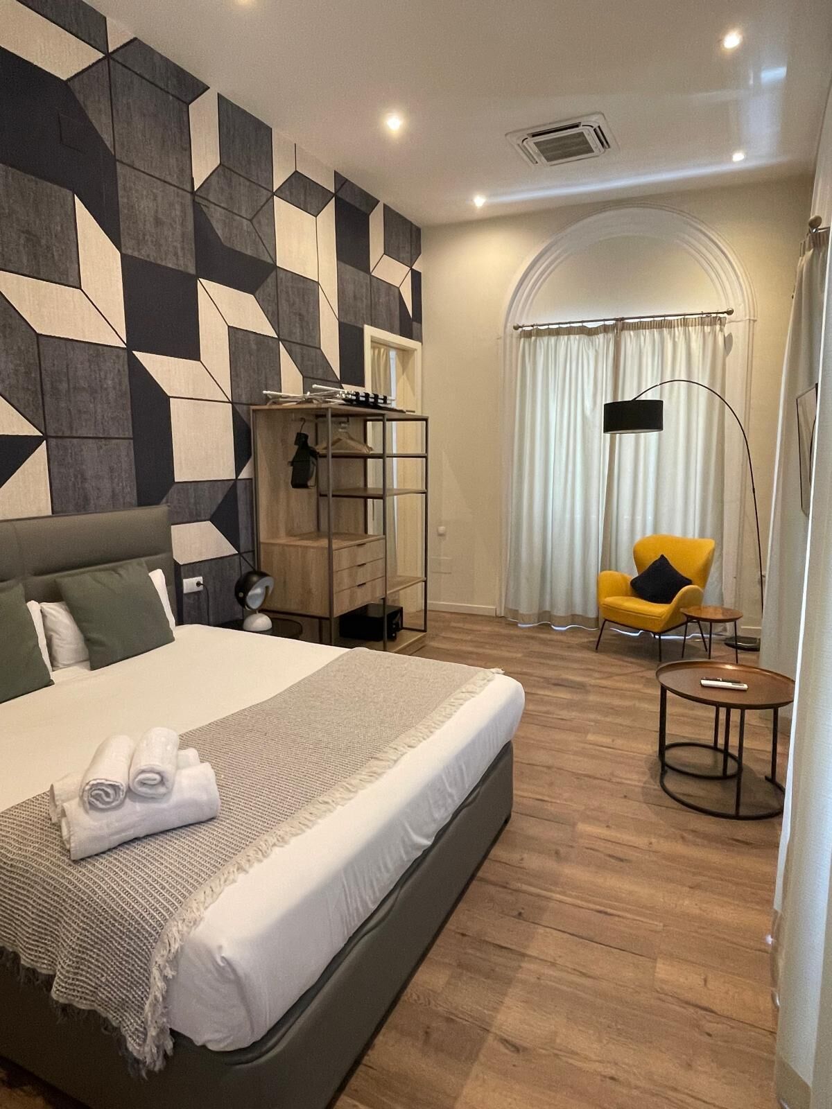 A modern hotel room with a large bed, a wardrobe, a yellow armchair, and a small round table.