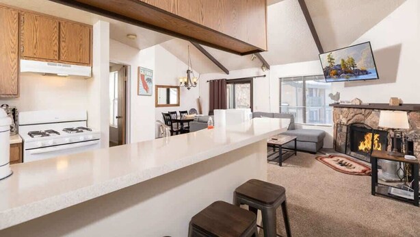 A modern kitchen with a white countertop, wooden cabinets, and a built-in stove. There is a dining area with a table and chairs, and a living room with a fireplace and a television mounted on the wall.