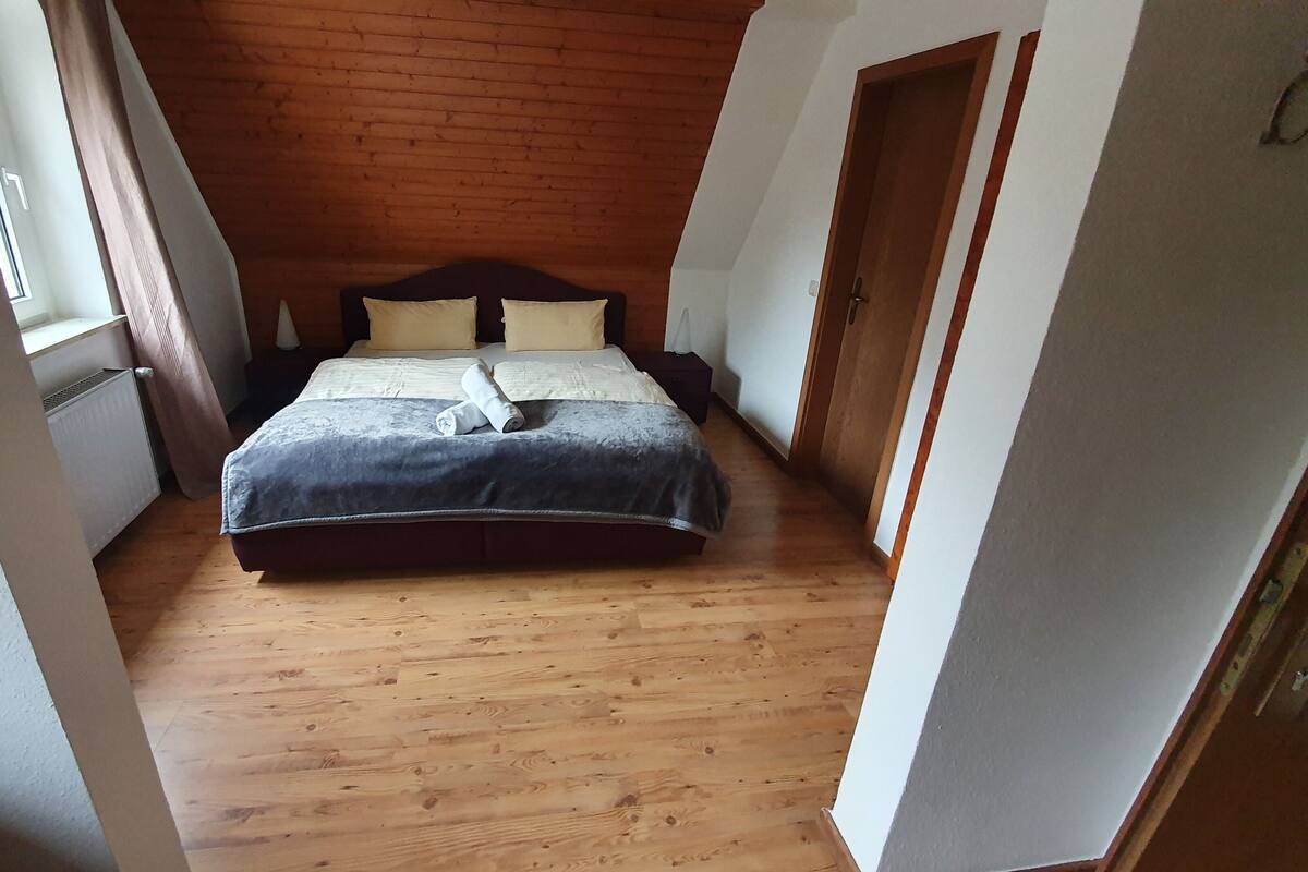 A bedroom with a wooden floor, a bed with a dark headboard, and a window on the left.