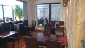 A restaurant with wooden tables and chairs, a potted plant, and a view of the outside through large windows. - Hotel Gartenstadt (Dortmund)