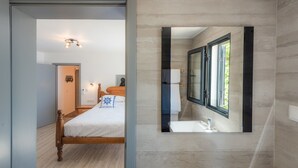 Une chambre moderne avec un lit en bois, une table de chevet et un miroir fixé au mur qui reflète une salle de bain avec un lavabo et une fenêtre.
