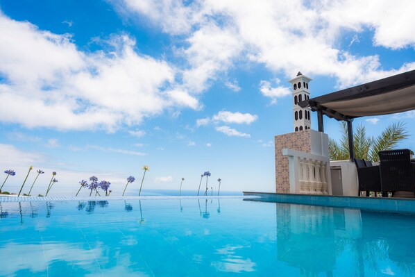 Une piscine avec une eau d’un bleu limpide, un bâtiment blanc surmonté d’une tour et un ciel dégagé.