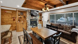 Un intérieur chaleureux avec une cheminée en pierre, des poutres apparentes en bois au plafond, un coin repas et un canapé placé près d’une fenêtre.