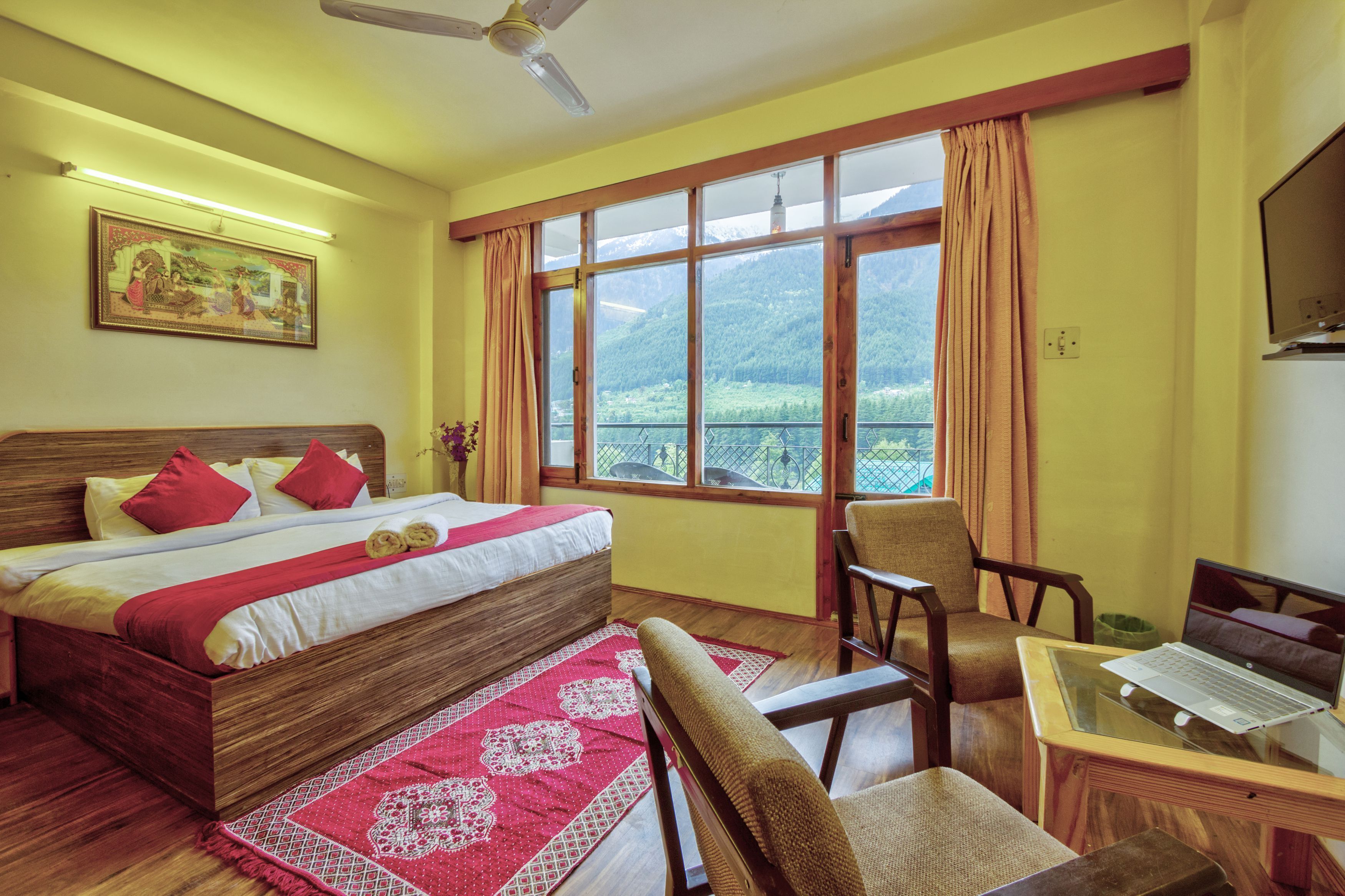 A hotel room with a bed, a desk with a laptop, a chair, a window with a mountain view, and a ceiling fan.