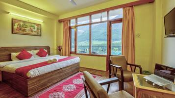 A hotel room with a bed, a desk with a laptop, a chair, a window with a mountain view, and a ceiling fan.