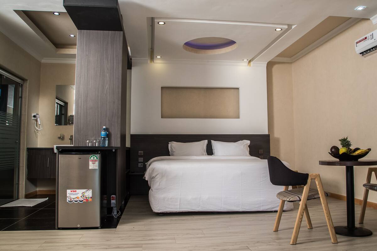 A hotel room with a bed, a desk with a chair, a refrigerator, and a small table with fruits.
