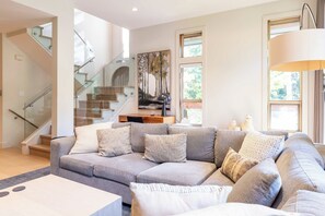 Una sala de estar moderna con un sofá gris, una mesa de centro, una escalera con barandillas de vidrio y un cuadro en la pared.