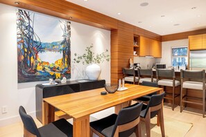 A modern kitchen with wooden cabinets, a dining area with a wooden table and chairs, and a large abstract painting on the wall.